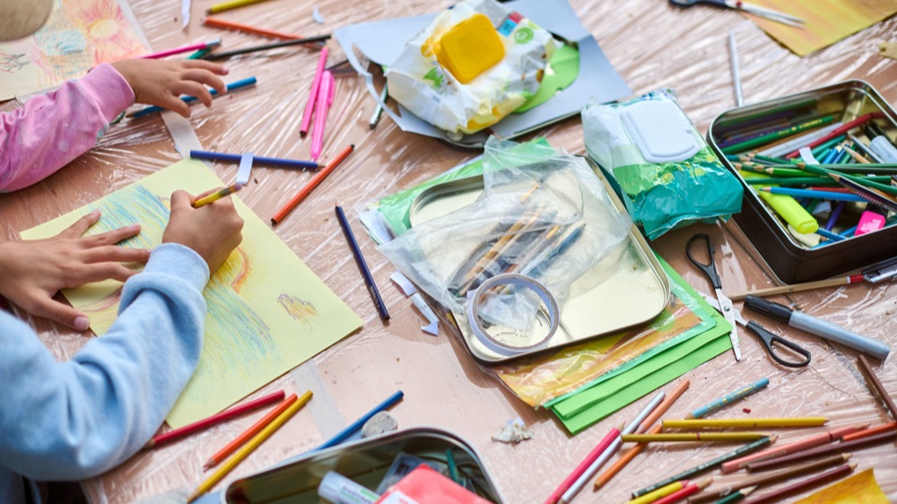 Kinderhände malen mit bunten Stiften an einem Tisch voller Bastelmaterial