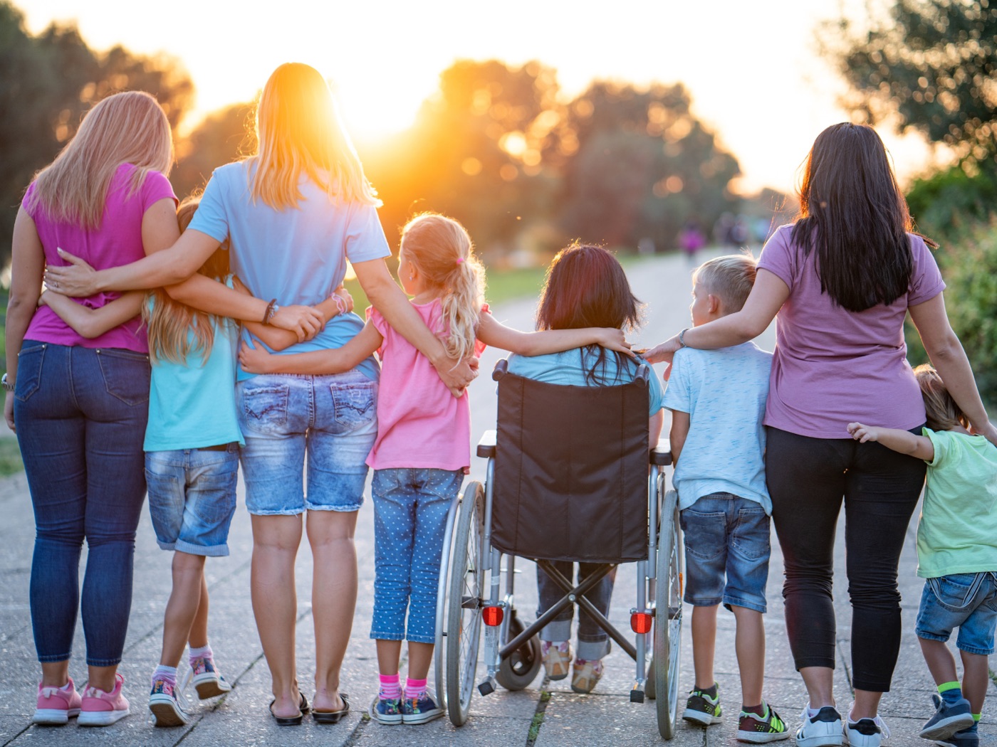 Kinder und Erwachsene gehen Arm in Arm einen Weg entlang, ein Kind sitzt im Rollstuhl